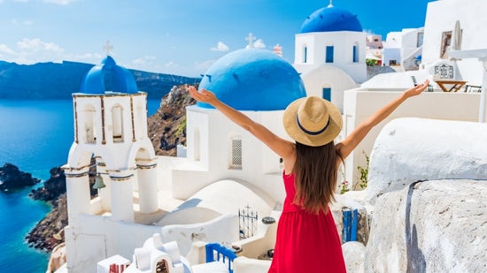 Person wearing a red dress and straw hat standing with arms outstretched among Santorini's whitewashed buildings and blue-domed churches, overlooking the Aegean Sea. 