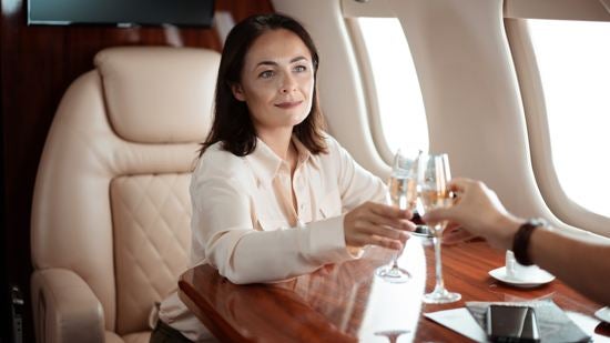 A woman on a private jet touches her champagne glass to a champagne glass held by the passenger opposite her.