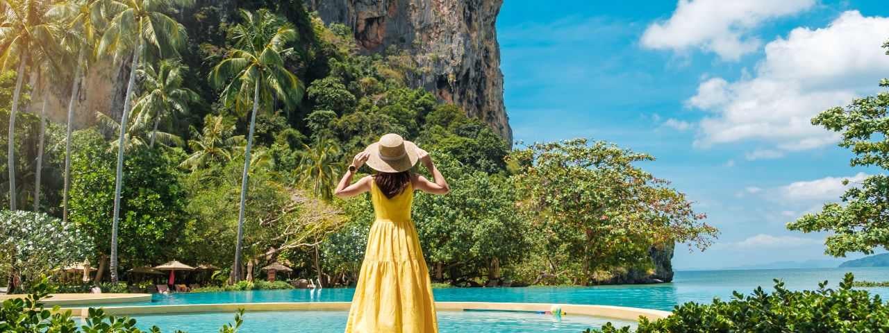 a woman tourist in a yellow dress and hat standing at a pool in front of the ocean at Railay Beach, Krabi in Thailand