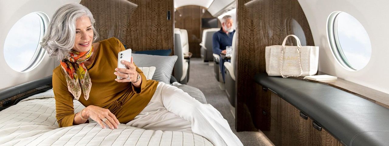 An attractive woman lies on her side on the bed in a Gulfstream G700 sleeping area while speaking on her phone.