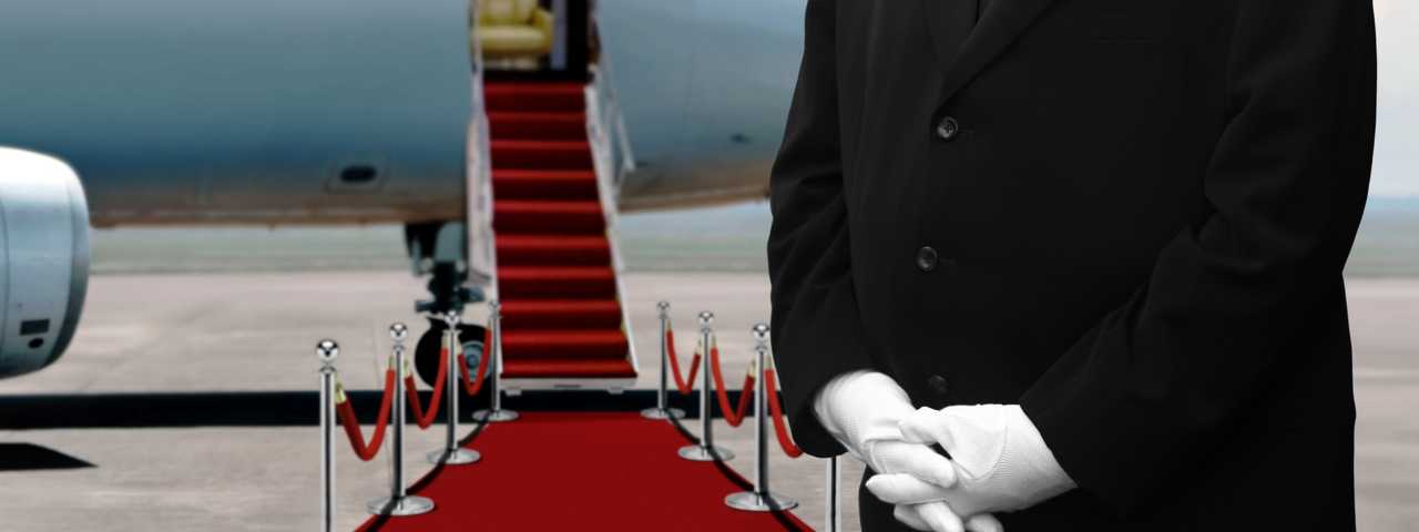 A man in a suit waits in front of a red carpet leading up into a private plane.