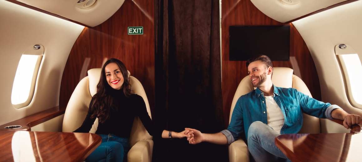 A young, smiling man and woman hold hands across the aisle in a private jet.