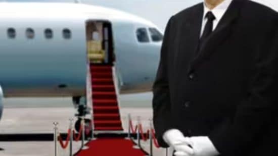 A white-gloved attendant waits at a roped red carpet that leads to the stairs of a private jet.