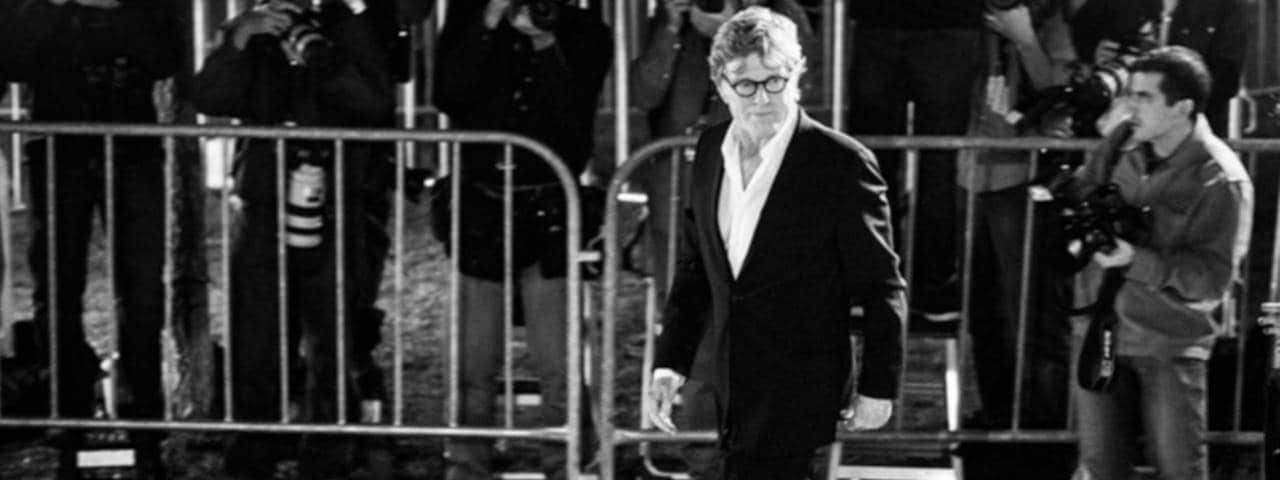 Black and white image of movie actor Robert Redford wearing glasses and a suit being photographed at an event