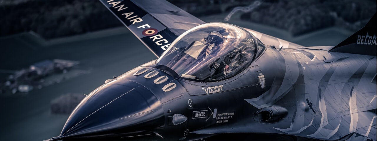 Ein Pilot sitzt im Cockpit eines eleganten, dunklen Kampfjets, der durch den Himmel fliegt.