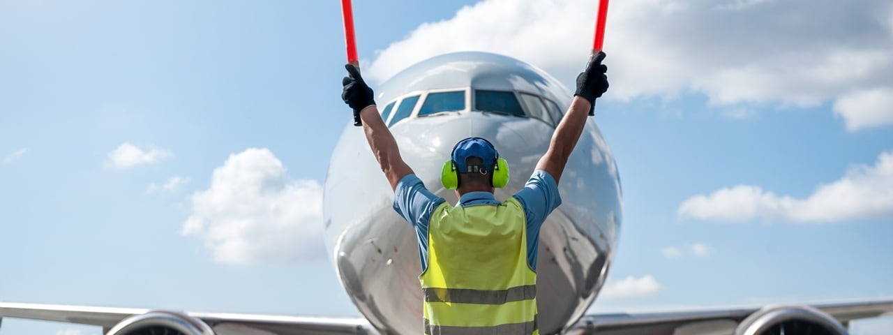 Ground crew in a signal vest holding signaling sticks in front of a commercial airliner.