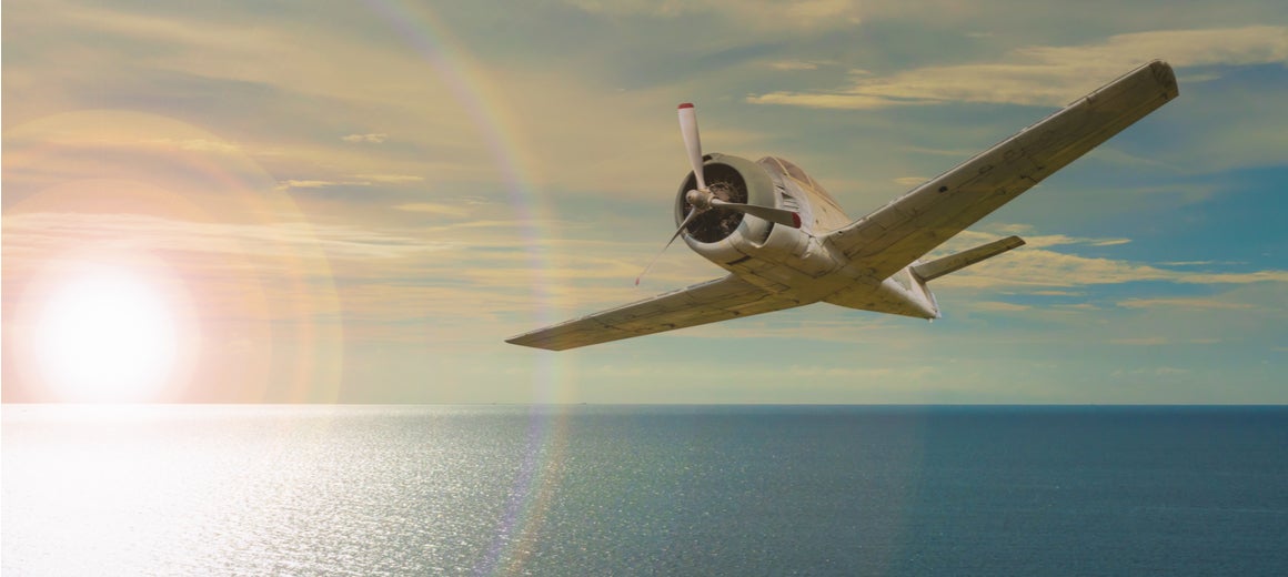 A vintage plane flies over the sea with the sun setting in the background.