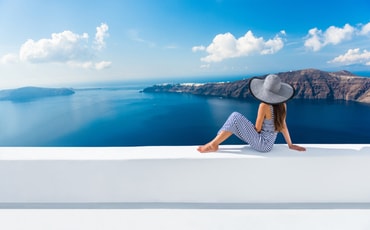 A woman in a sunhat sitting on a white wall looks out over the ocean and distant mountains