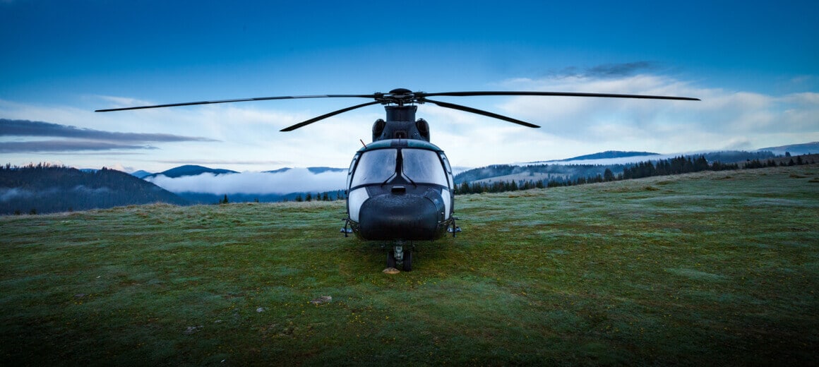 A private helicopter parked on a field with a forest, mountains and clouds in the background.