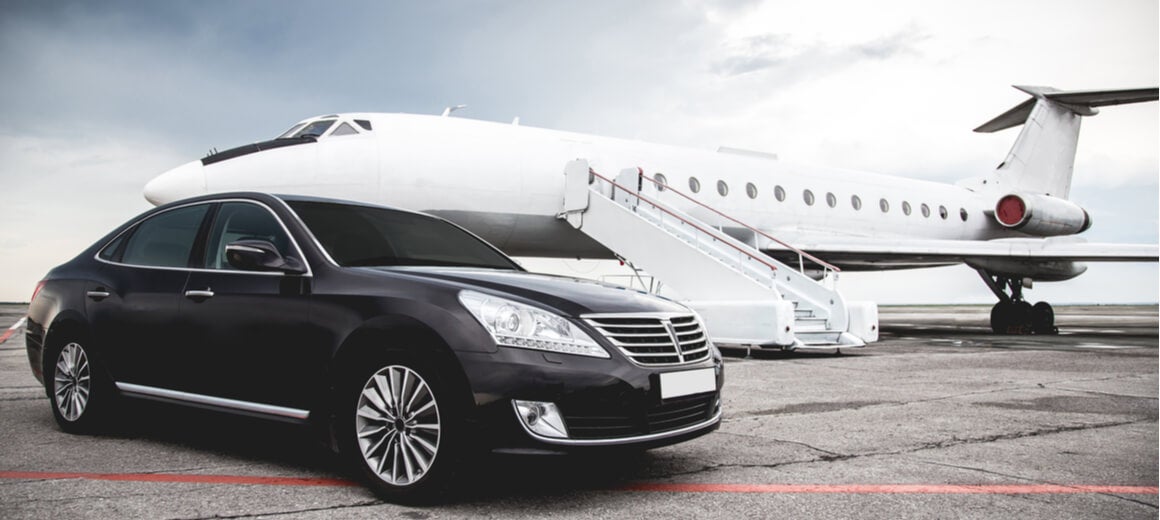 A private jet and a black luxury car stand parked on a runway in overcast weather.