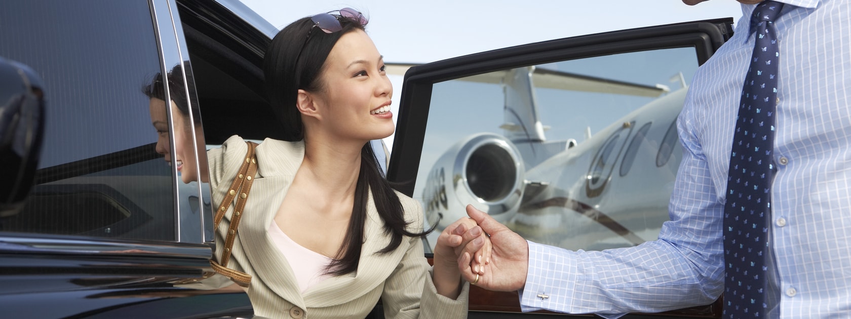 A man in a suit helps a well-dressed woman get out of a car with a private jet in the background.