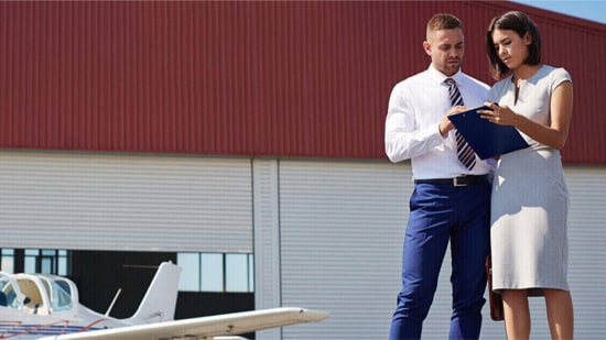 A smartly dressed man and woman look at a clipboard outside an aircraft hangar.