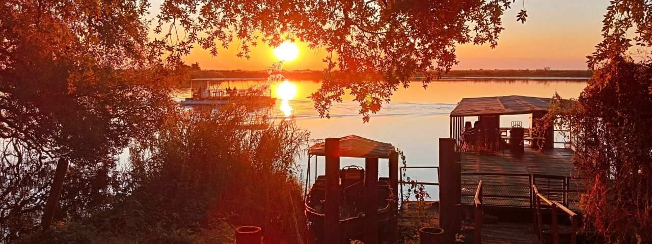 A photograph of a sunset over the Chobe River in Botswana. A boat sits on the land, and another is floating on the river.