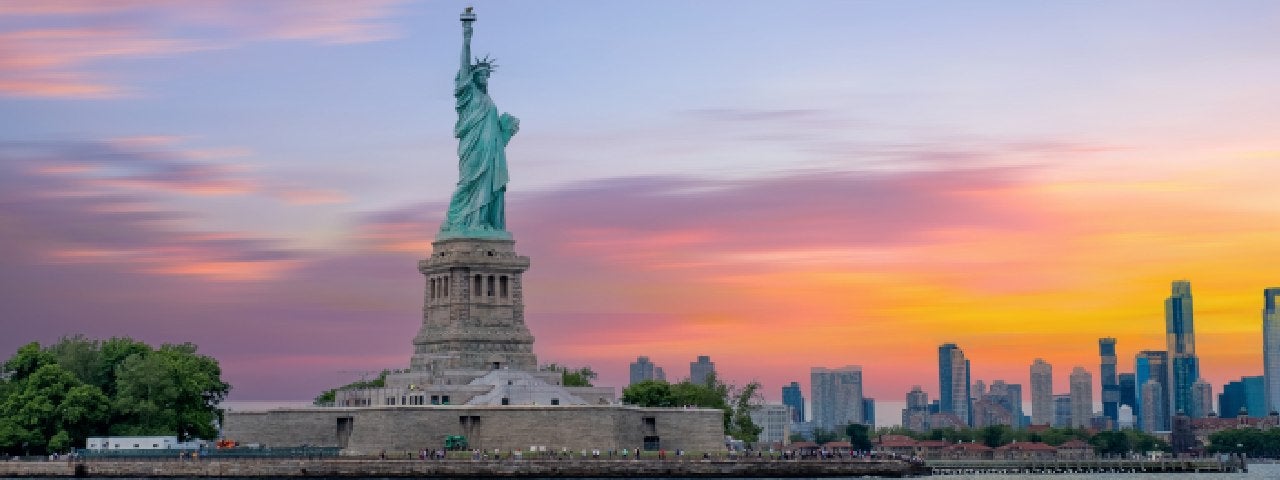 A photograph of the Statue of Liberty with the New York skyline in the distance, at sunset.
