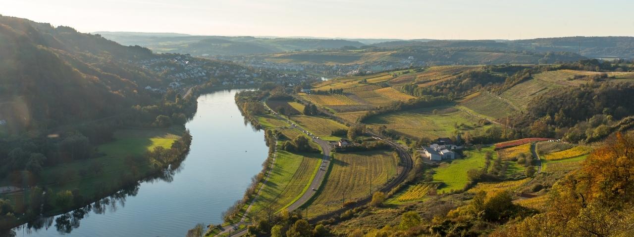 Aerial view of a winding river flowing through a broad valley surrounded by patchwork agricultural fields, vineyards, roads and scattered buildings. Rolling hills and elevated terrain extend into the distance under soft daylight. 