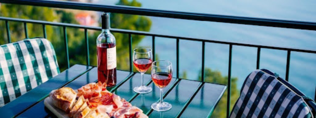 Outdoor table set with sliced cured meats, bread, and two glasses of red wine overlooking a body of water.