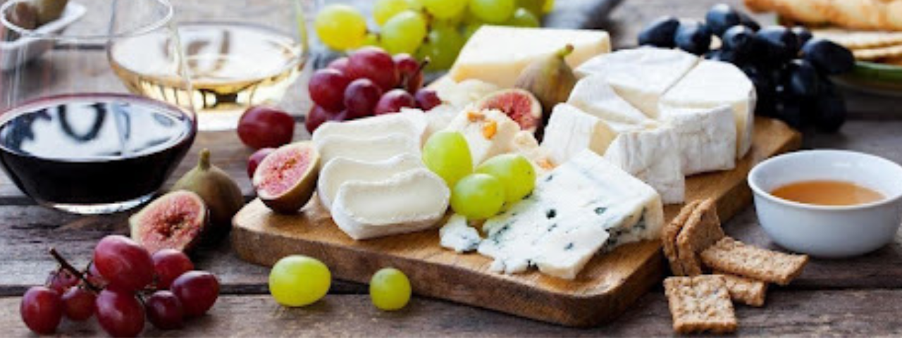Cheese board with assorted soft and blue cheeses, grapes, figs, crackers, and glasses of red and white wine on a wooden table.