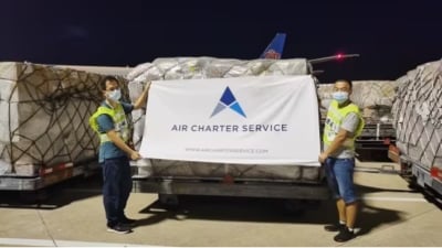 Two men wearing high visibility vests and masks and holding up an Air Charter Service banner in front of cargo pallets.