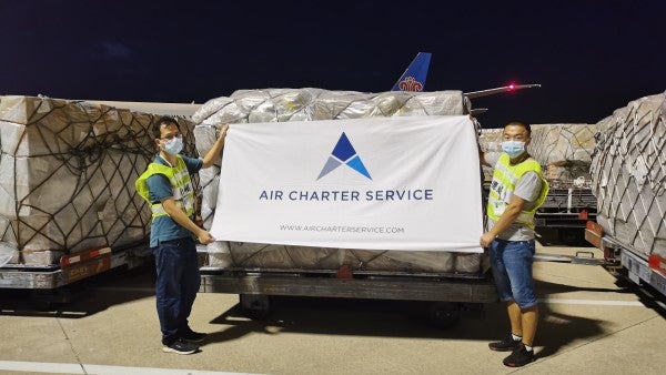 Two airport staff holding up an Air Charter Service banner in front of cargo palettes about to be loaded onto a cargo plane.