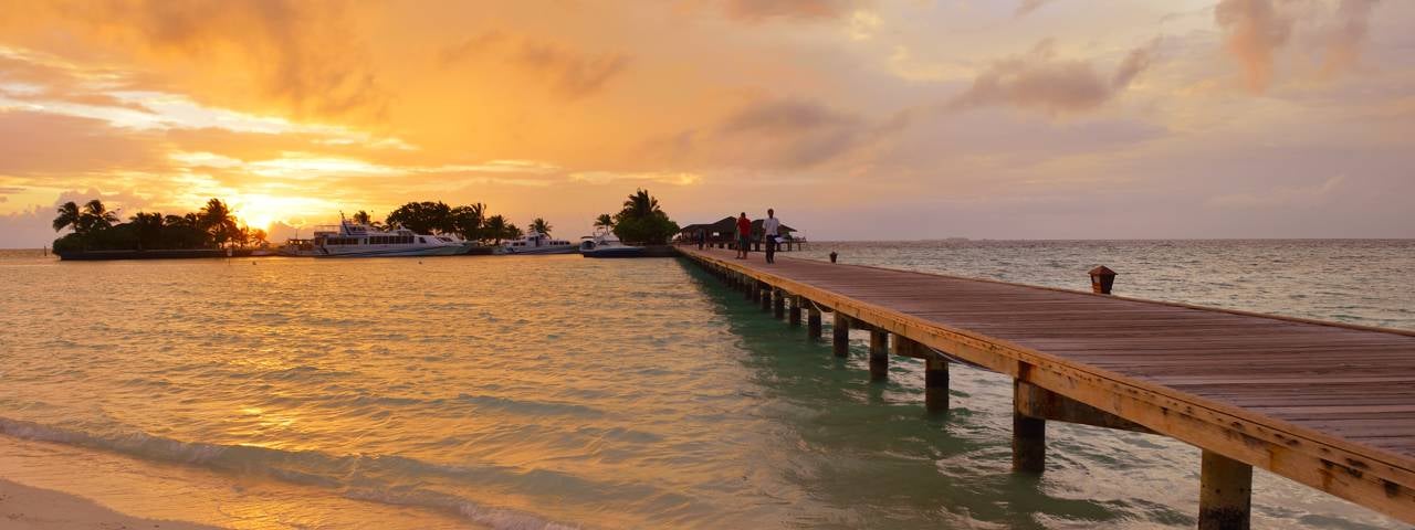 A long wooden pier extends over calm turquoise water toward several boats docked at a tropical island under a vibrant orange and yellow sunset.