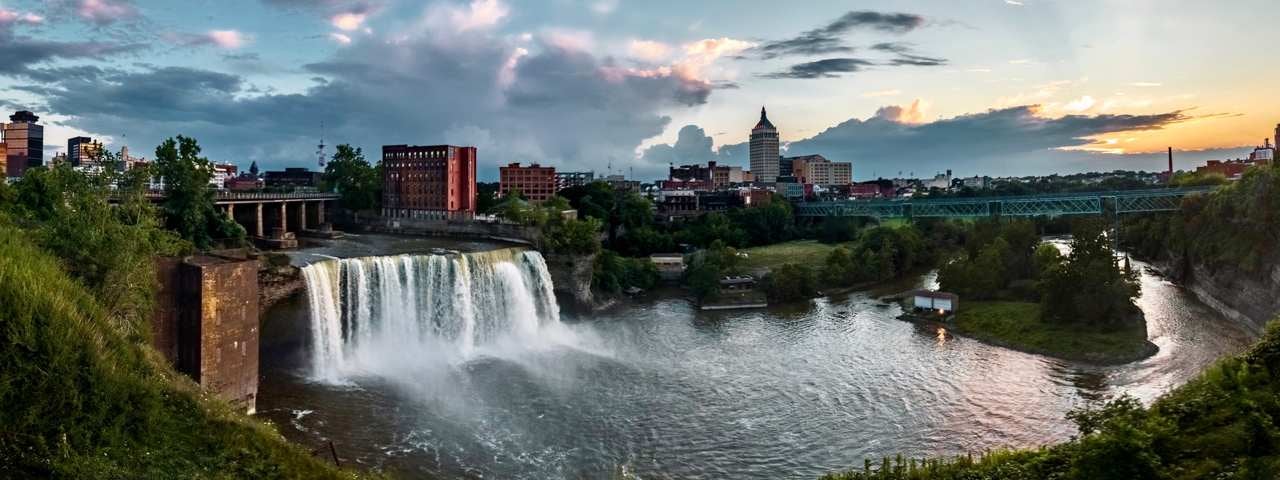 A small waterfall and river with Rochester skyline in the background at sunset