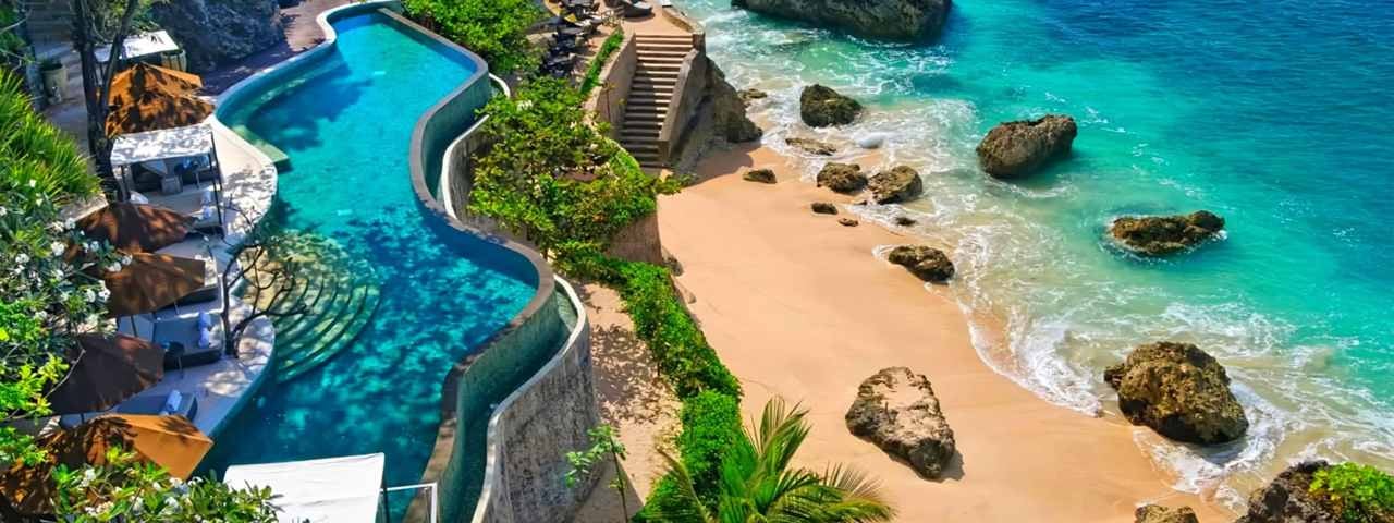 A stunning aerial view of a luxurious resort featuring a curvy infinity pool adjacent to a sandy beach with turquoise waters. The pool area is surrounded by lush greenery and several sun loungers with brown umbrellas. Stone steps lead down to the beach, which is dotted with large rocks and boulders.