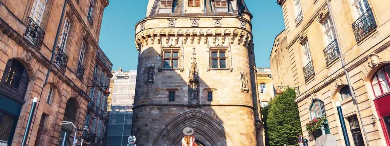 A woman standing in front of the Porte Cailhau in Bordeaux on a sunny day.
