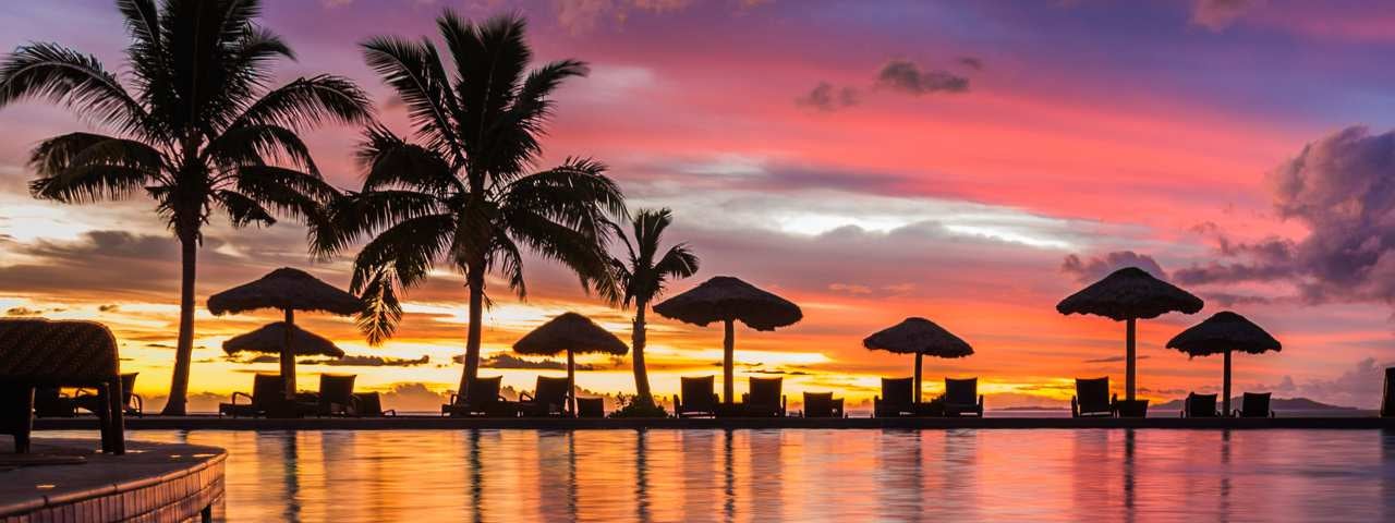 A sunset view from a swimming pool onto the beach with palm trees and thatch beach umbrellas.