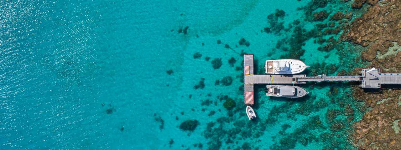 An aerial view of two luxury boats parked next to a dock jutting out into the turquoise ocean in Kadavu, Fiji.