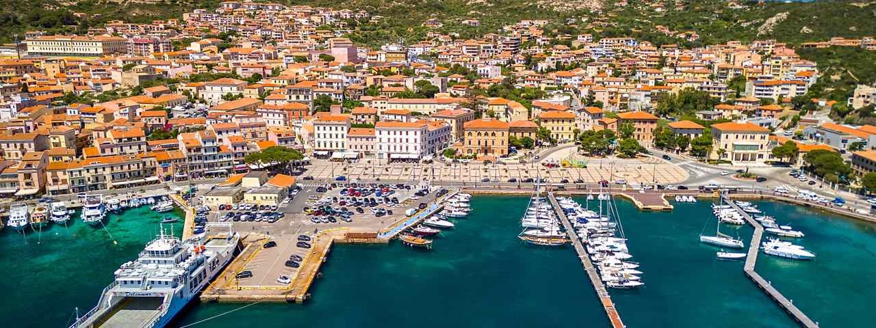 A picturesque seaside town in Sardinia, with luxury yachts and a ferry docked in the harbour.