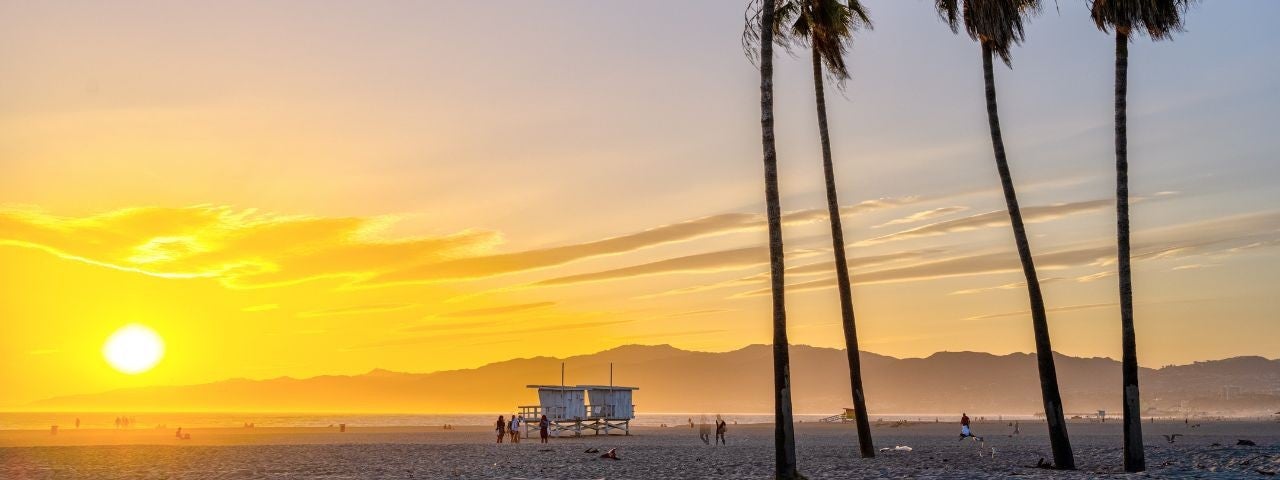 Sunset at Venice Beach in Los Angeles, with tall palm trees, a lifeguard tower and people walking along the sandy beach, against a backdrop of mountains and a glowing sky.