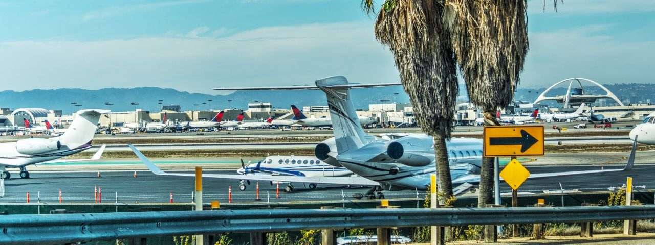 View of private jets and commercial airplanes on the tarmac at Los Angeles International Airport (LAX), with palm trees and the airport’s iconic Theme Building in the background.