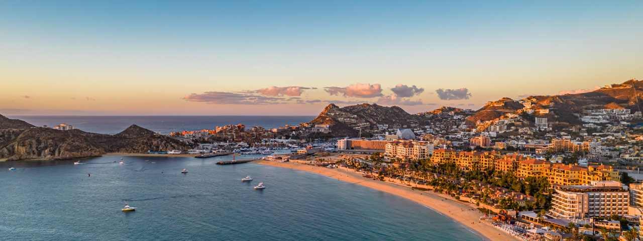 A sunset beach scene in Cabo San Lucas with boats out on the ocean and buildings and hills to the side. 