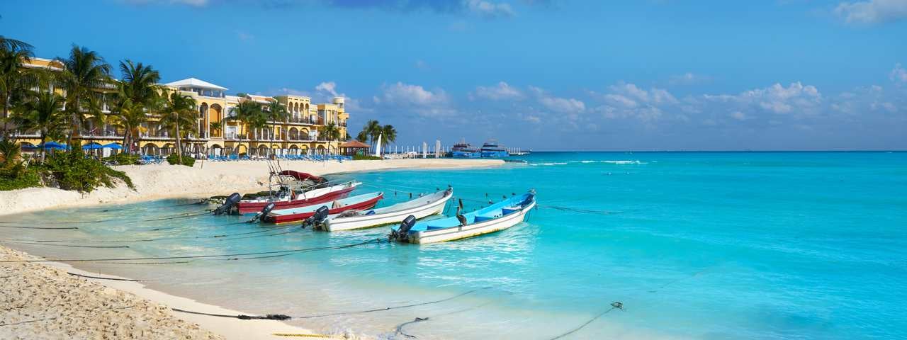 Boats parked just off of a stunning beach in Playa del Carmen with a hotel and palm trees in the background