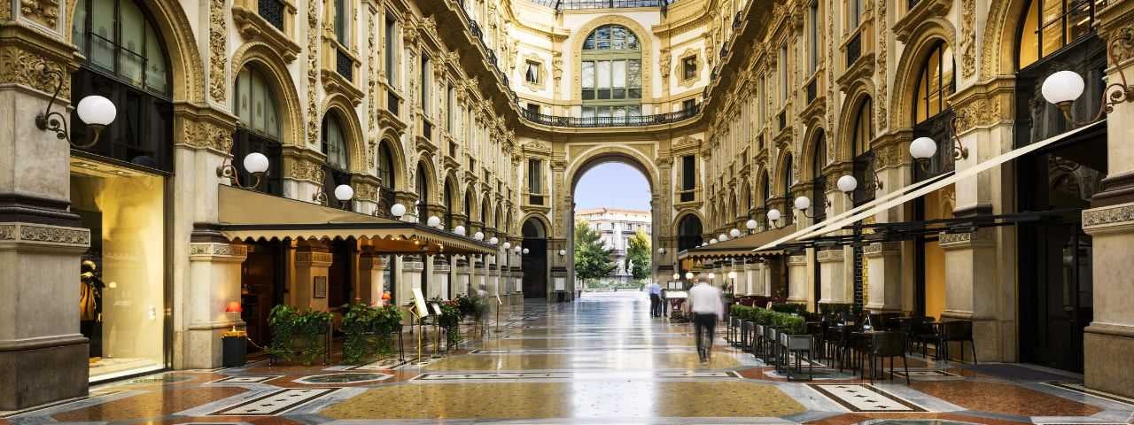 Elegant interior of Galleria Vittorio Emanuele II in Milan, Italy, featuring ornate architecture, mosaic floors, and outdoor seating for cafés beneath arched walkways.