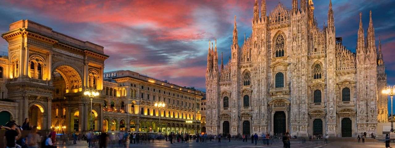 Evening view of the Milan Cathedral (Duomo di Milano) and the illuminated Galleria Vittorio Emanuele II entrance in Piazza del Duomo under a dramatic sunset sky.