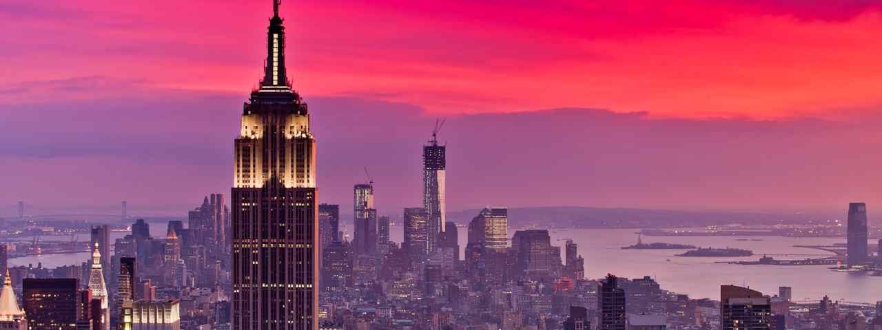 An aerial view of the New York City skyline with the ocean in the background and purple and red sunset hues in the sky.