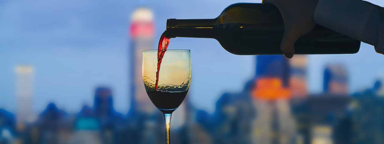 A person pouring a glass of red wine in the evening with the New York City skyline in the background.