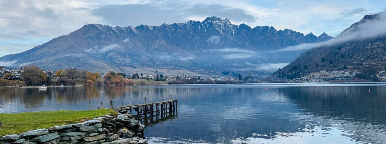 A weathered wooden jetty juts out over Lake Wakatipu, its calm waters backed by the Remarkables mountain range.