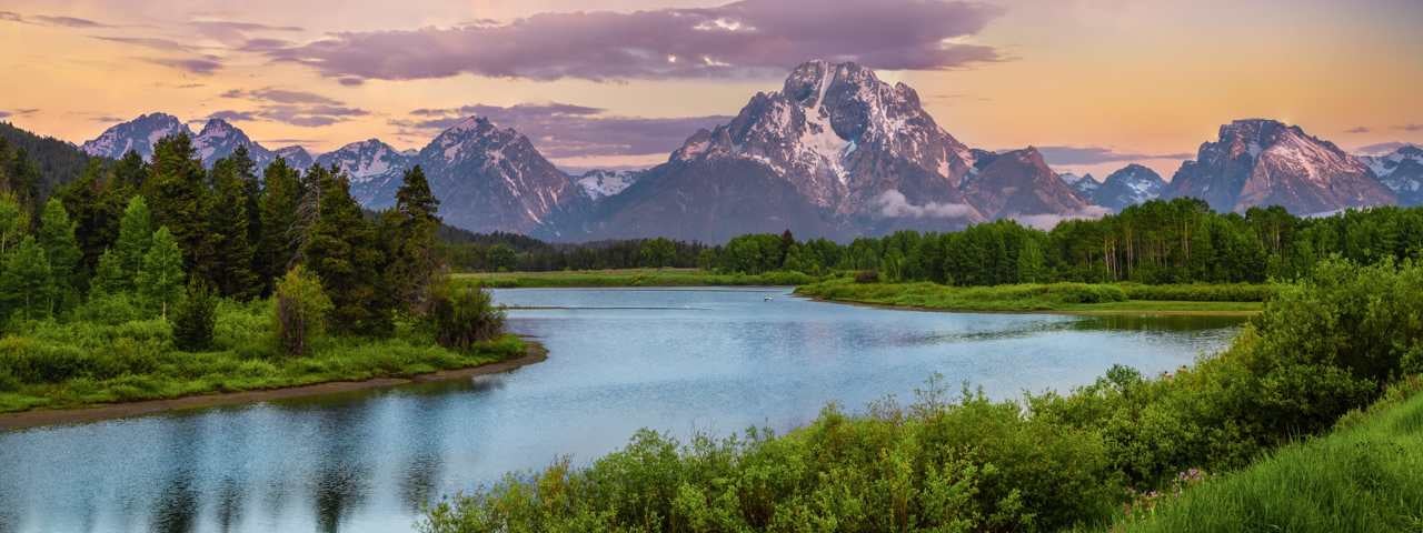 A lake surrounded by trees and mountains with the sunset behind them in Wyoming