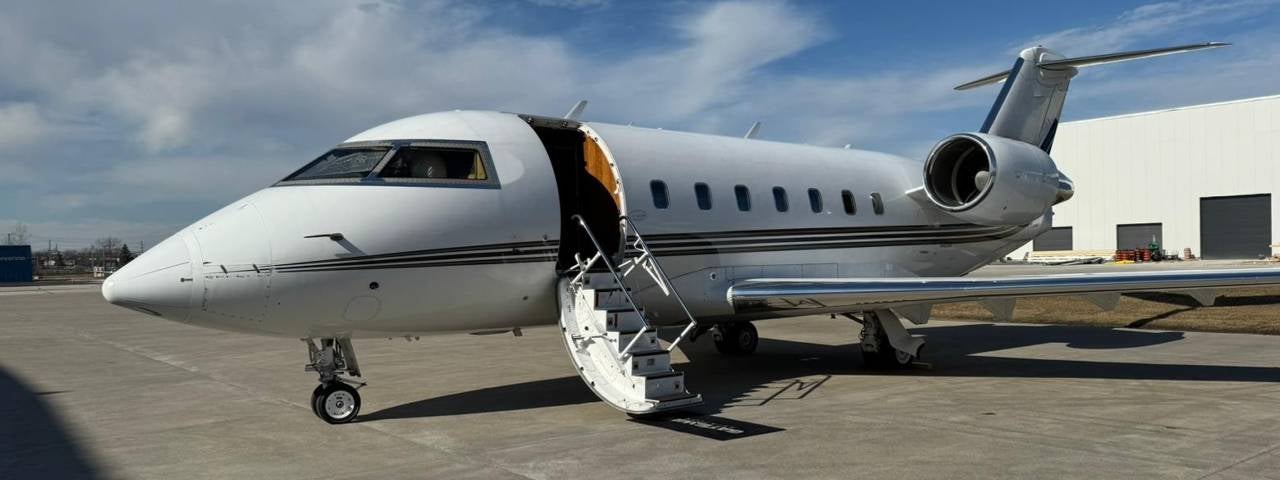 Sleek white private jet on sunny airport tarmac; cabin door open and curved stairs lowered, hangar building behind