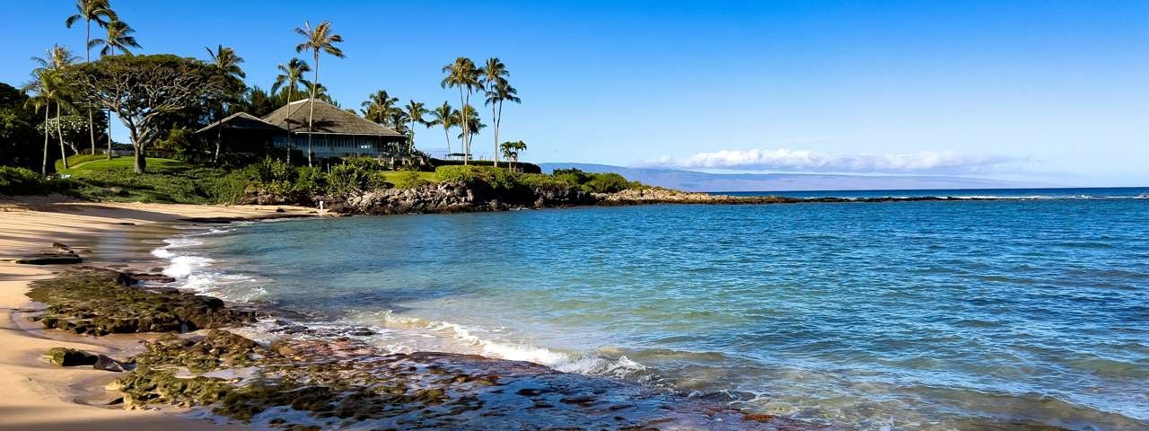 A beautiful beach on a sunny day in Maui with palm trees and a hotel in the background.