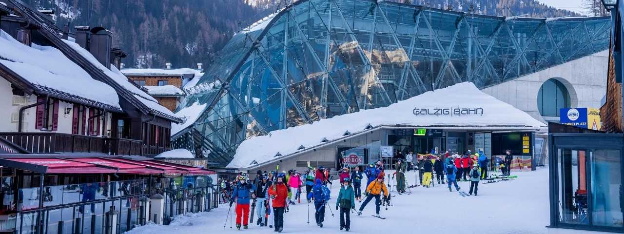 Skiers gathered outside a modern glass cable car station surrounded by snow-covered buildings, with colorful winter clothing adding contrast to the alpine setting. Visitors carrying skis and poles make their way across the snowy plaza beneath the striking curved structure. Nearby chalets and mountain slopes create a lively après-ski atmosphere. 