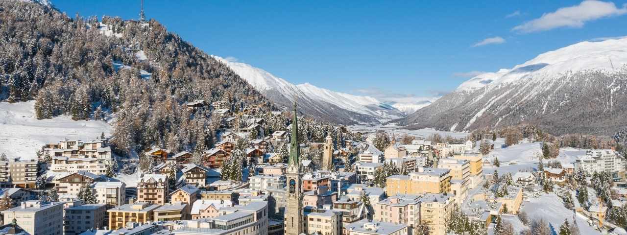 Panoramic view of a snow-covered alpine town with church spires, pastel buildings, and forested mountains rising above a broad valley. Sunlight illuminates the rooftops and surrounding peaks, revealing layers of snow-covered hills in the distance. The mix of historic architecture and dramatic scenery gives the town a classic alpine character. 