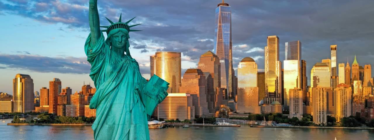 The Statue of Liberty stands in the foreground with the New York City skyline glowing behind it at sunset.