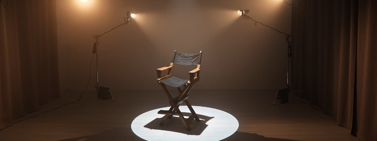 An empty black wooden director's chair positioned in the centre of a stage spotlight on a light wood plank floor, flanked by two vintage studio lights on tall metal stands with dark curtains draping the background in a dimly lit production set. 