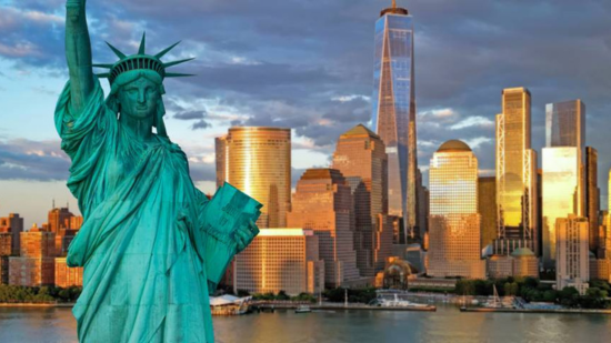 The Statue of Liberty stands in the foreground with the New York City skyline glowing behind it at sunset.