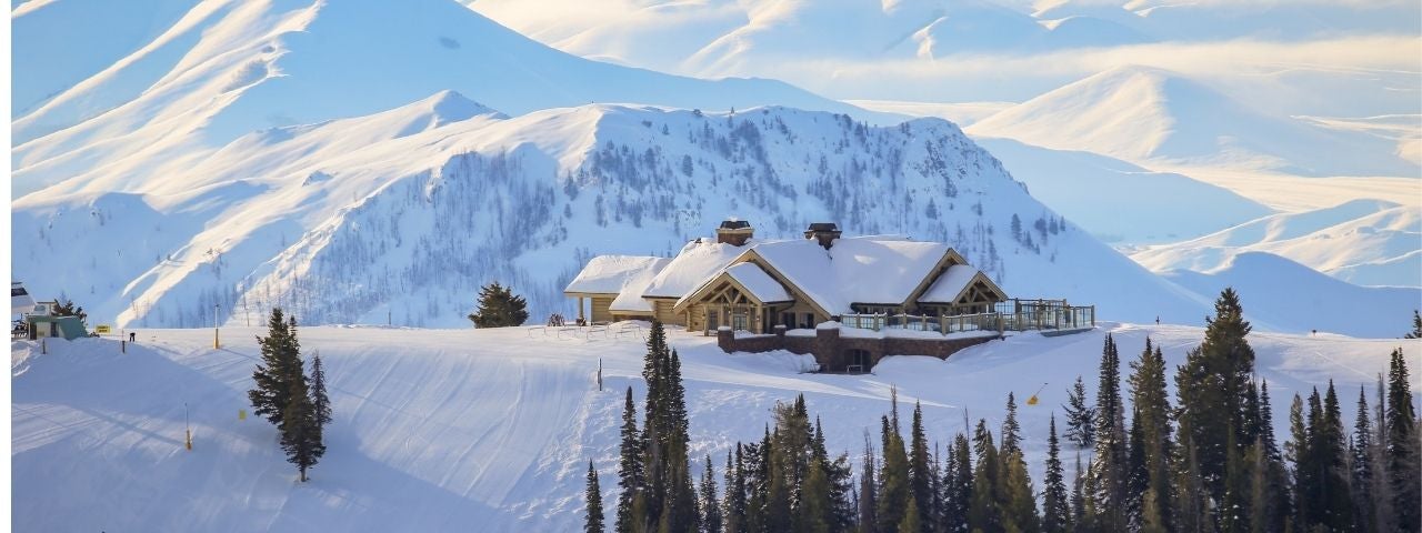 Snow-covered mountain lodge with a pitched roof sits on a ridge surrounded by evergreen trees. Rolling white mountain peaks extend into the distance beneath a clear blue sky, with deep snow covering the surrounding landscape.