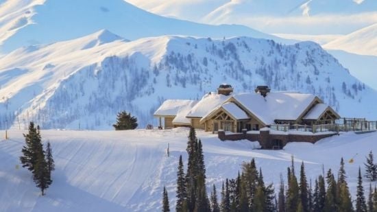 Snow-covered mountain lodge with a pitched roof sits on a ridge surrounded by evergreen trees. Rolling white mountain peaks extend into the distance beneath a clear blue sky, with deep snow covering the surrounding landscape. 