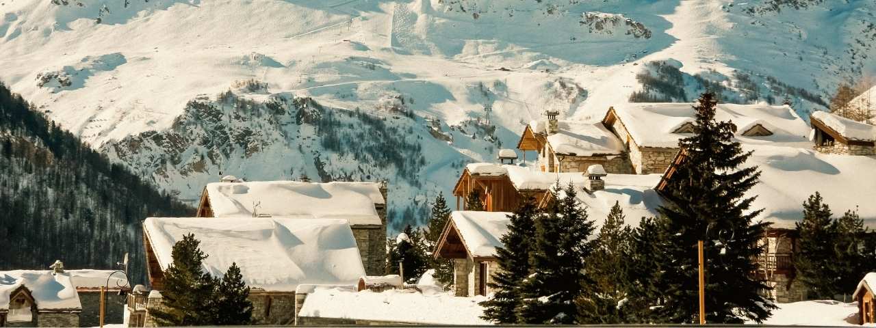 Traditional stone and timber chalets covered in deep snow overlooking expansive alpine slopes, with evergreen trees and rugged mountain scenery in the background. Layers of snowy rooftops and tall pines frame the elevated resort village nestled among the peaks. Bright winter sunlight casts soft shadows across the landscape, emphasizing the depth of the surrounding mountains. 
